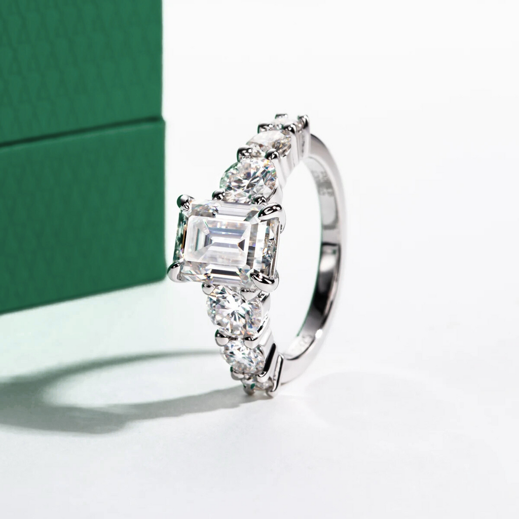 Anel prateado com moissanite central retangular e pedras arredondadas ao redor, fundo branco e caixa verde