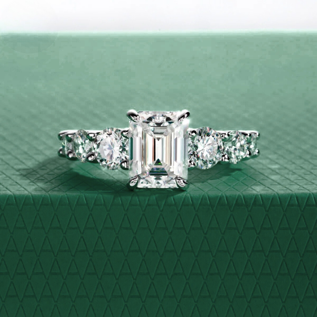 Anel Amara com pedra central moissanite de corte esmeralda e pedras laterais redondas, sobre fundo verde texturizado