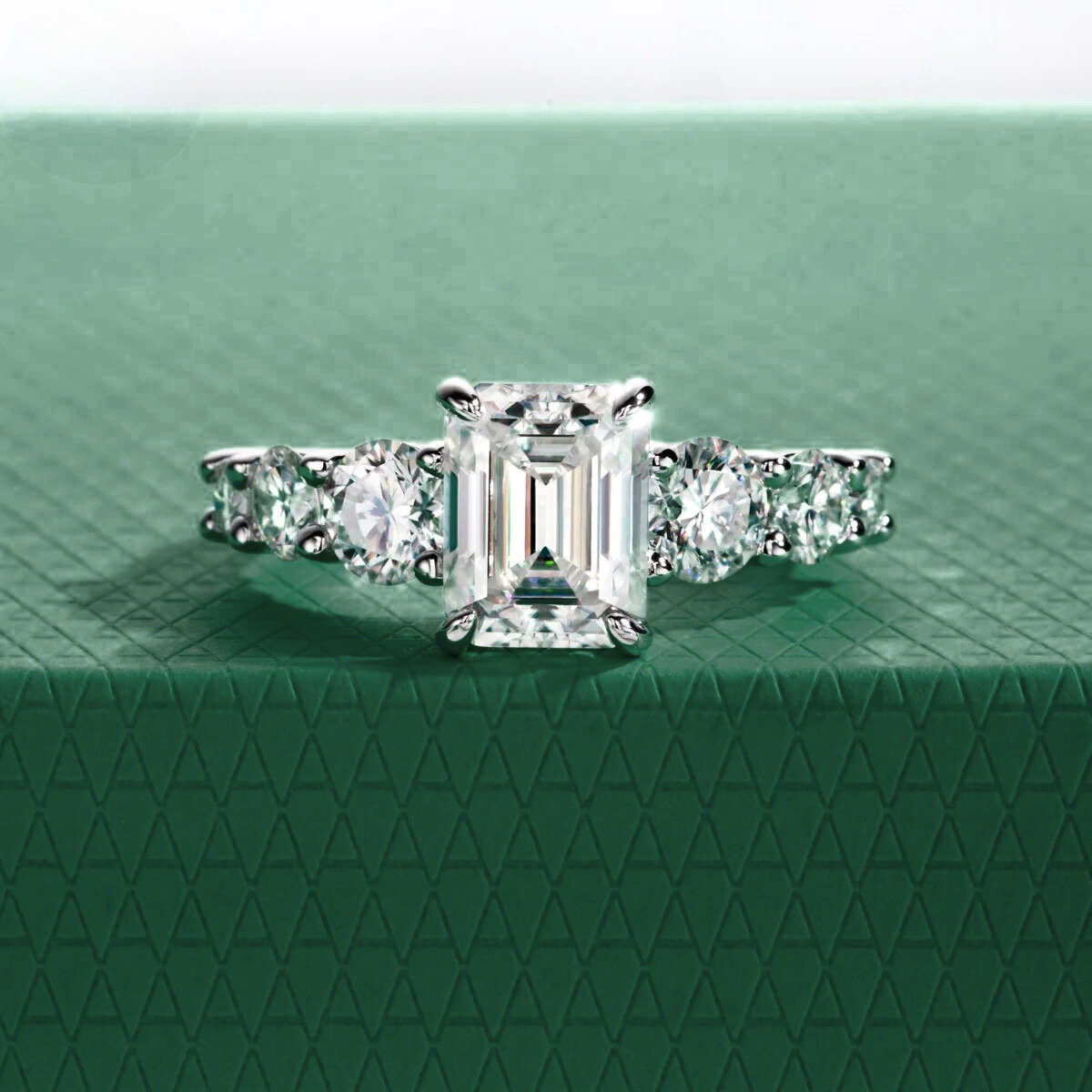 Anel Amara com pedra central moissanite de corte esmeralda e pedras laterais redondas, sobre fundo verde texturizado