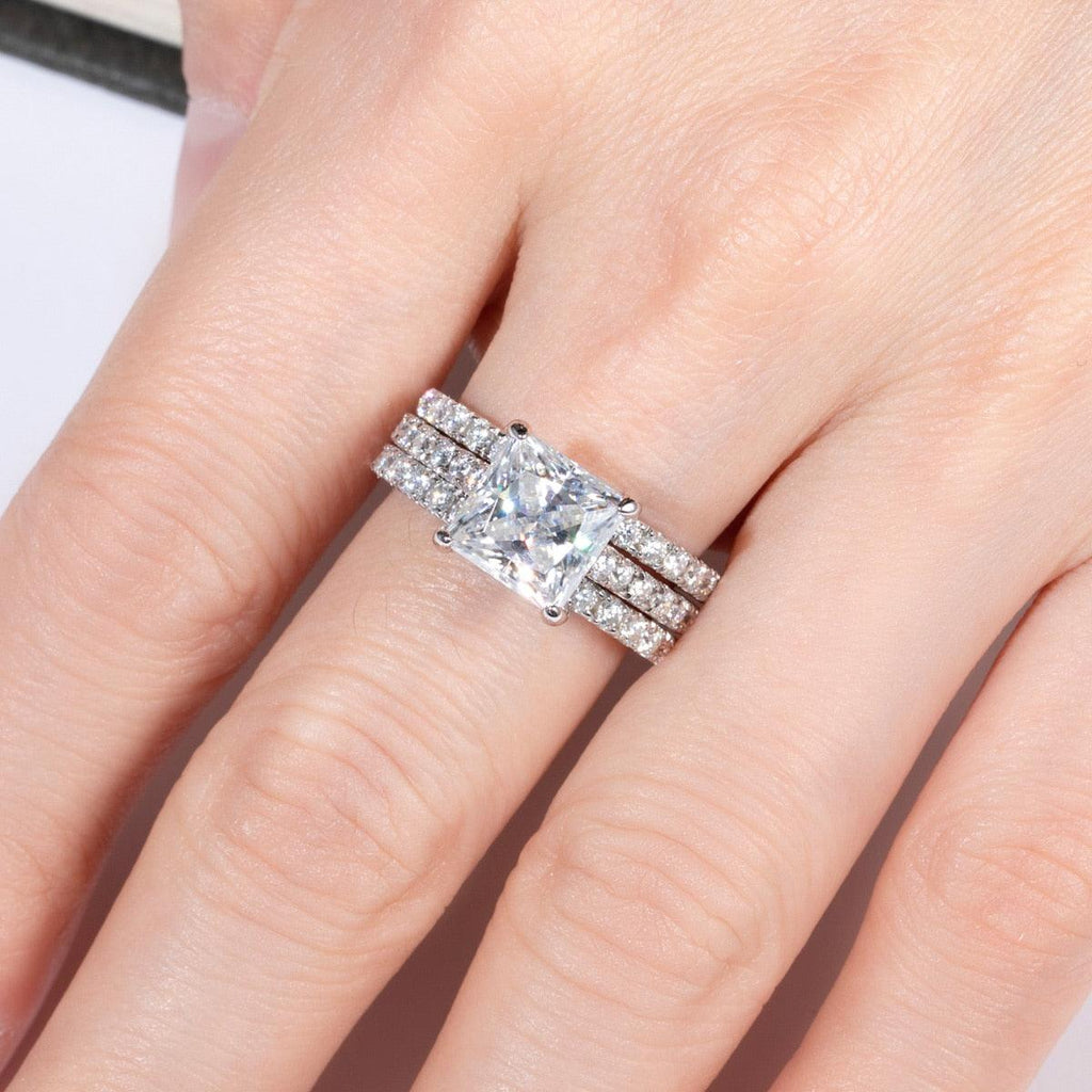 Anel delicado com pedra central quadrada de moissanite e faixas laterais cravejadas com pedras brilhantes, em dedo feminino